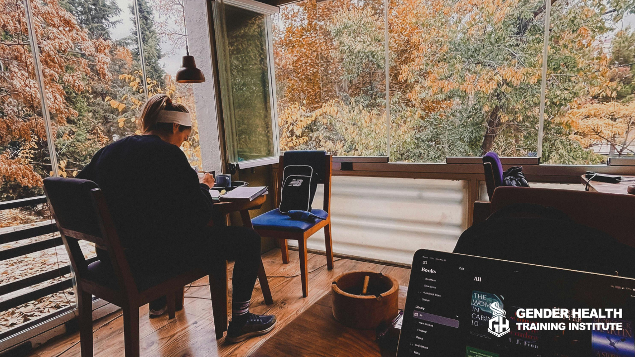 Professional cozying up to a laptop at a kitchen table in a room paneled by windows overlooking an autumnal forest while the professional enjoys LGBTQ+ Cultural Competency Trainings for CEU